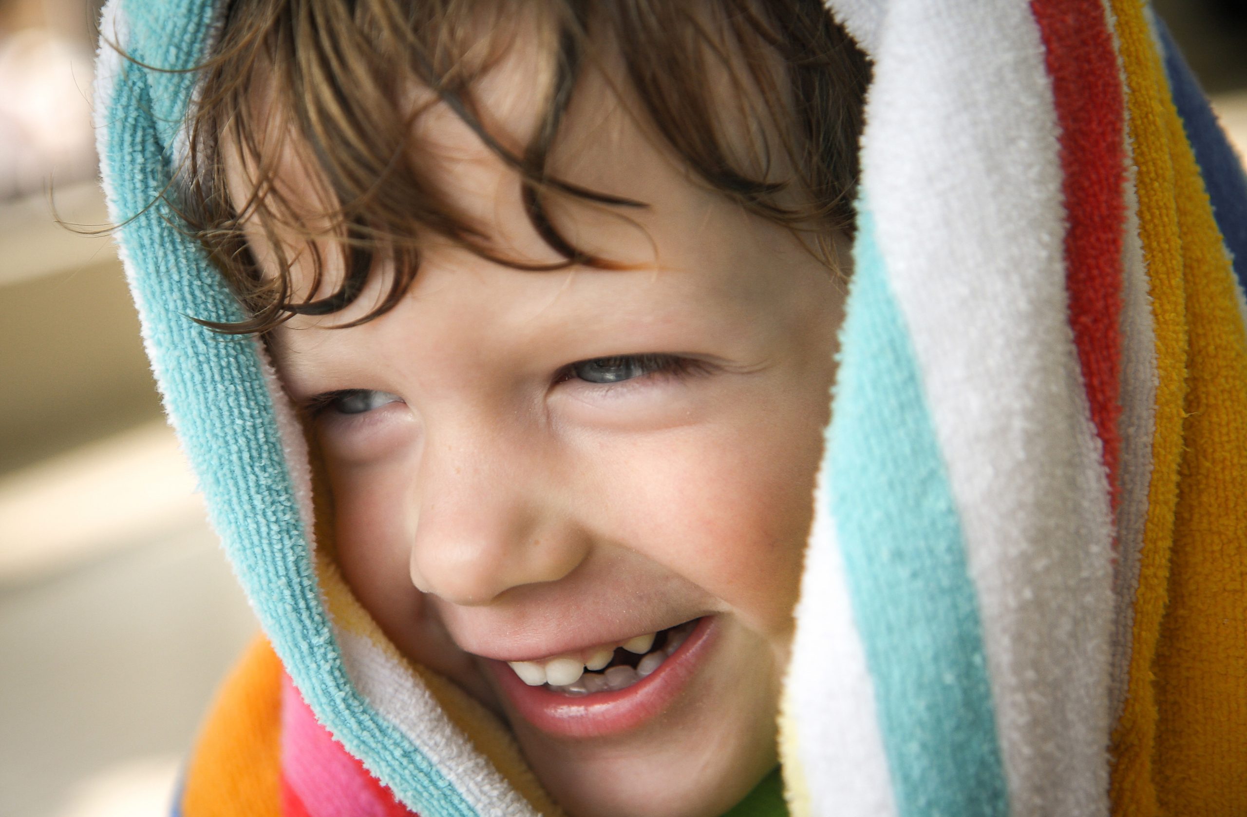 A young boy with a towel wrapped around his head smiles while drying off after getting out of a pool.